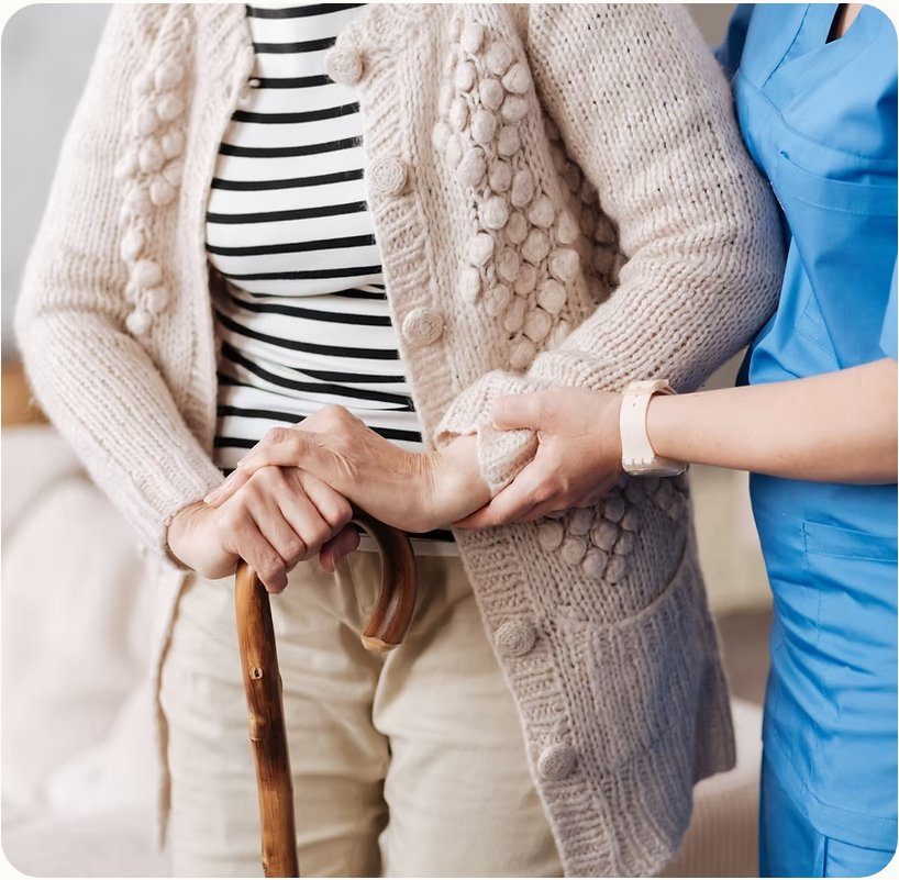 A nurse in blue scrubs gently supporting the arm of an elderly person using a wooden walking cane